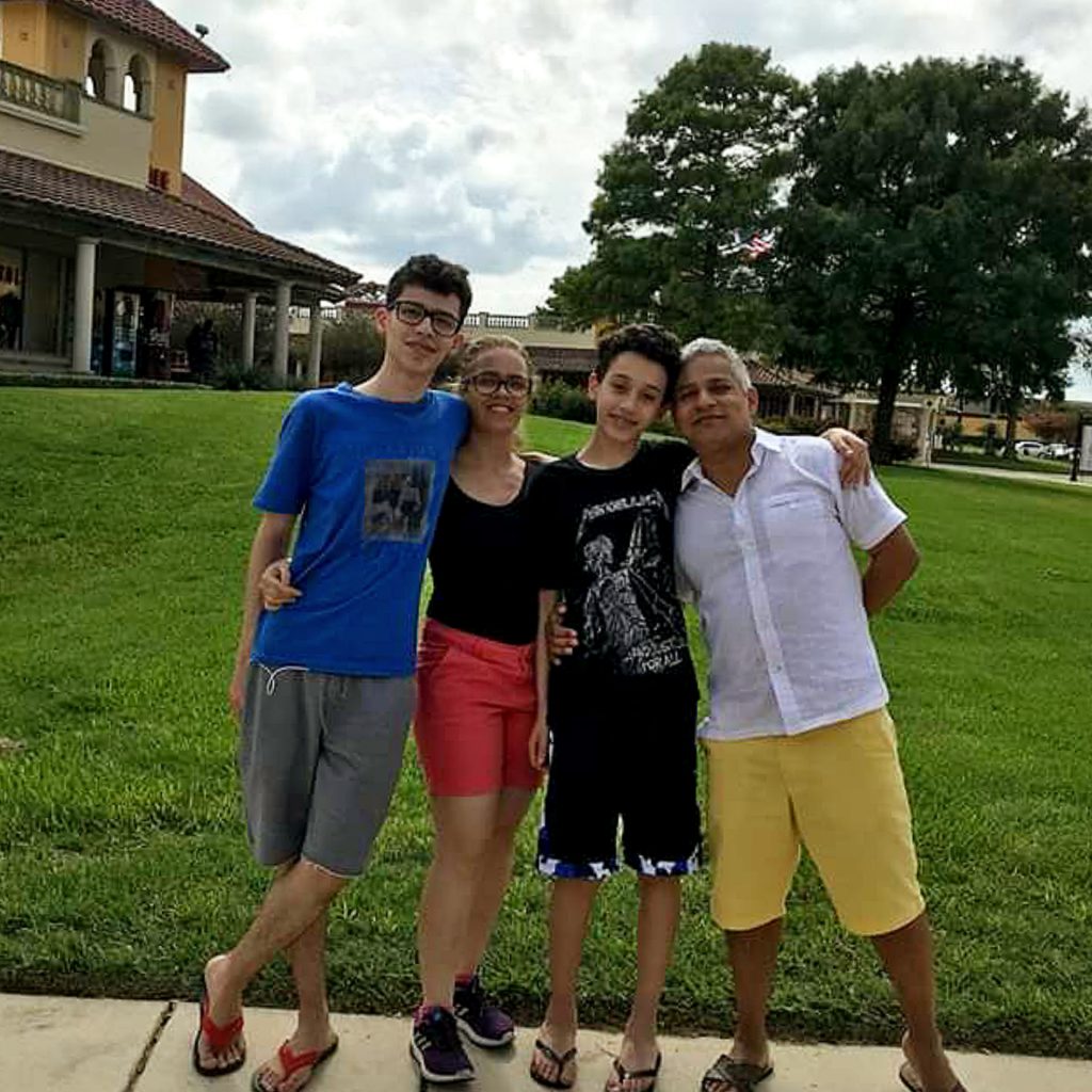 Casal e dois meninos adolescentes posam sorridentes em campo gramado. Ao fundo há uma pousada e árvores.