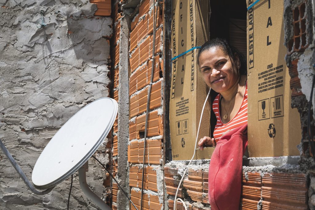 Mulher apoiada na janela de casa. Fachada com tijolos e reboco aparentes e do lado esquerdo, antena de tv a cabo.